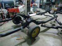Black car chassis on a garage floor with two wheels attached and other parts scattered about.