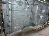 Underside of a car chassis, painted gray, held up on a rack in a workshop setting.