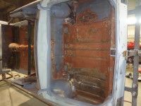 Rust-covered car chassis interior on a metal stand, possibly undergoing restoration; visible walls and floor.