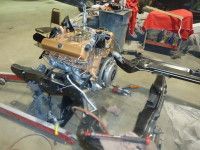 Engine being lowered into a black car frame in a garage. Gold and silver engine with surrounding tools.
