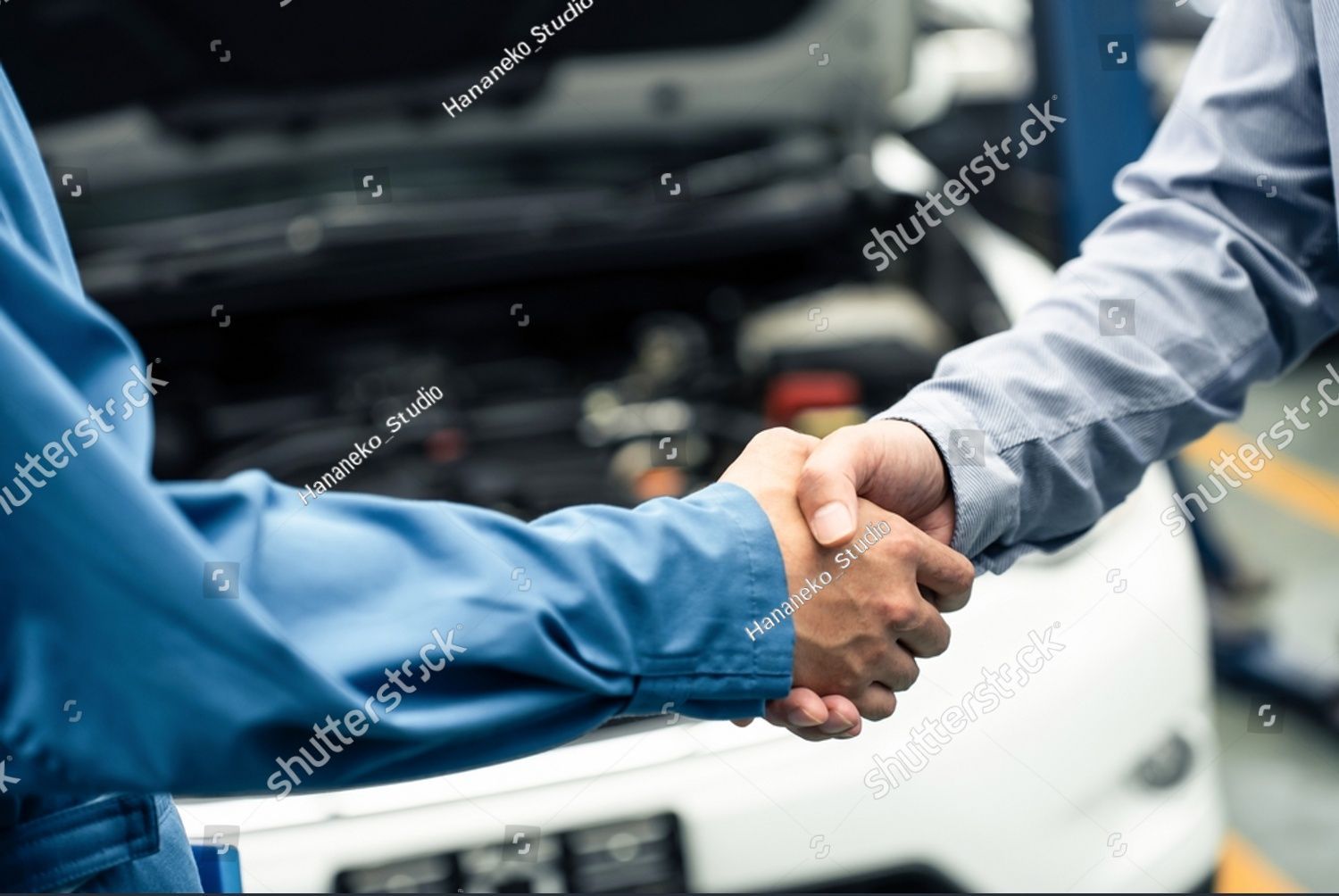 Two mechanics are shaking hands in a garage in front of a car.