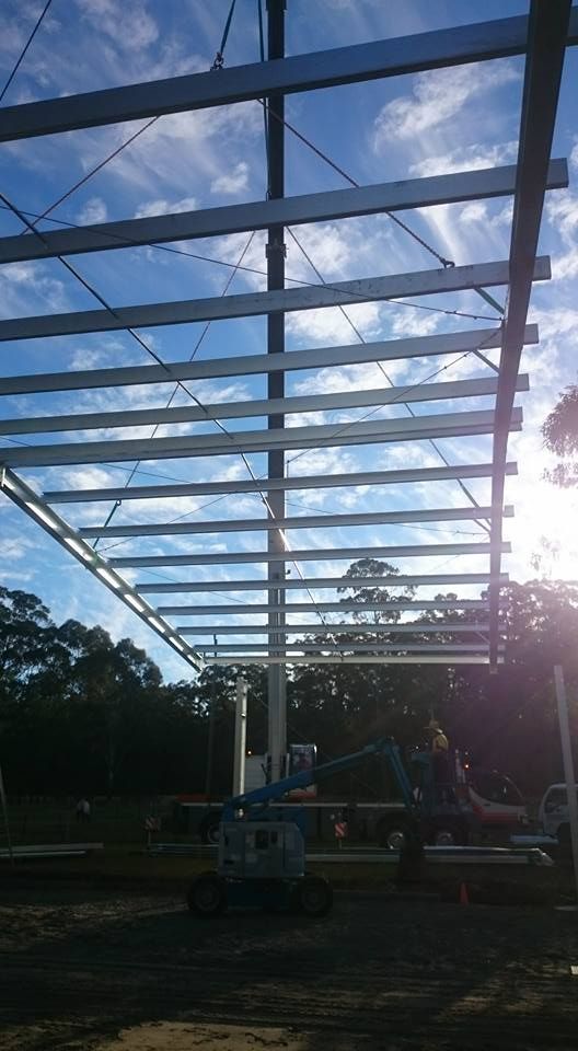 A Roof Steel Frame Structure Being Placed on posts by crane cables with the blue sky in the background — Burke Metal Fabrication In Taree, NSW