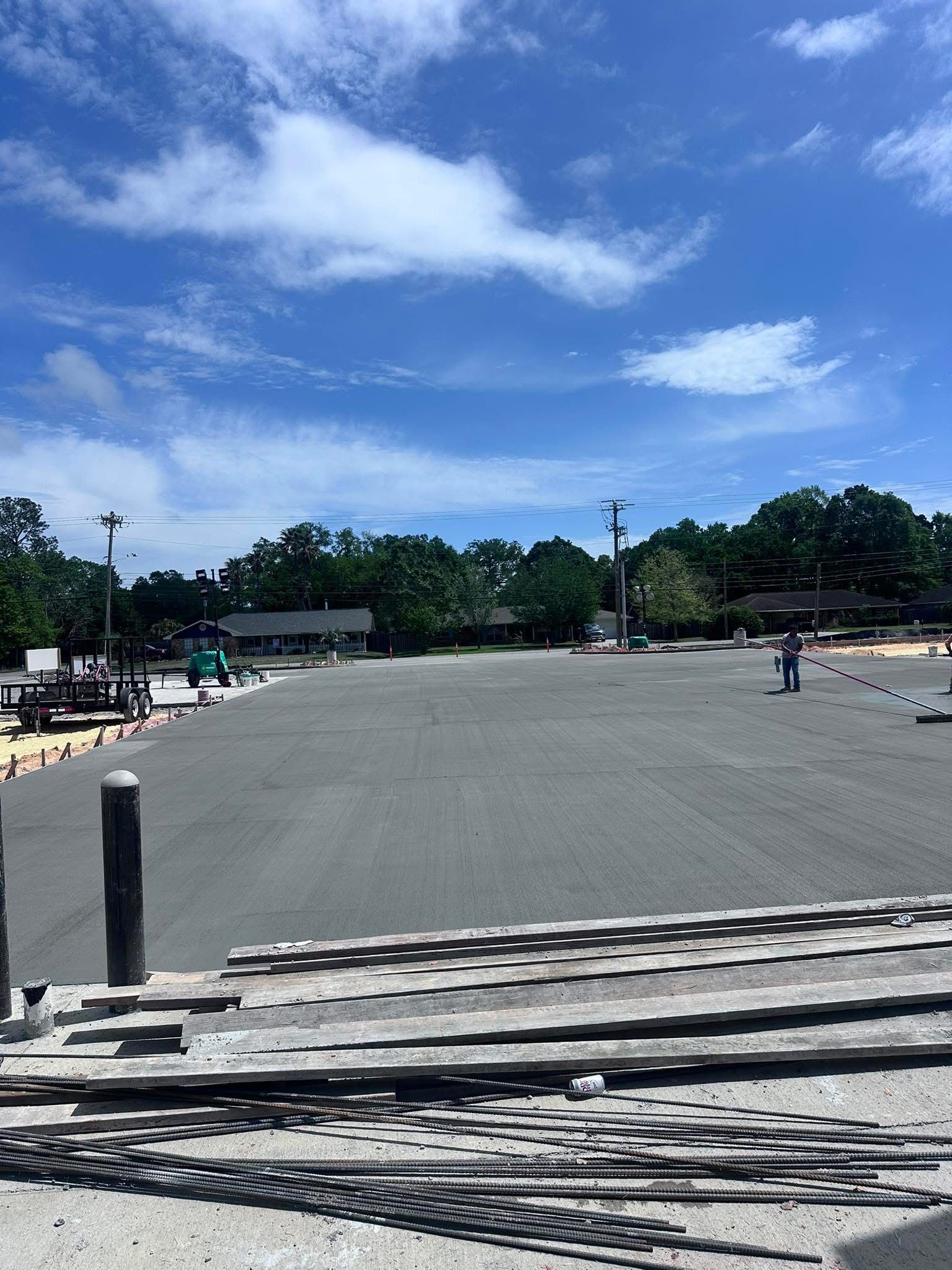 Wide empty paved lot under a bright blue sky with scattered clouds, trees and parked vehicles in the distance