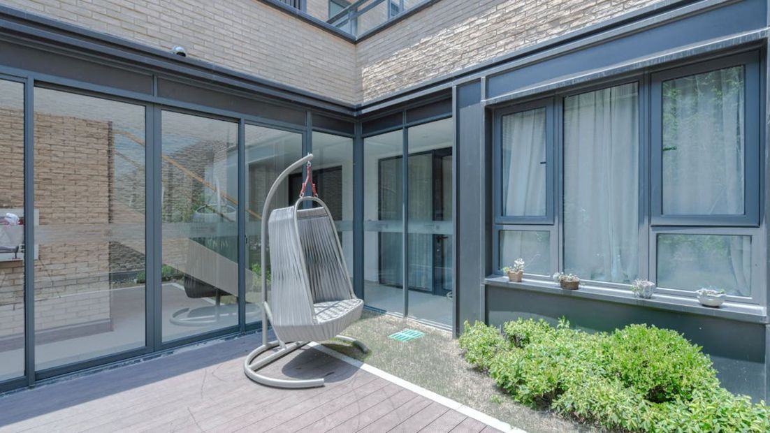 Patio moderno con paredes de cristal, una silla colgante y vegetación junto a un edificio de ladrillo.