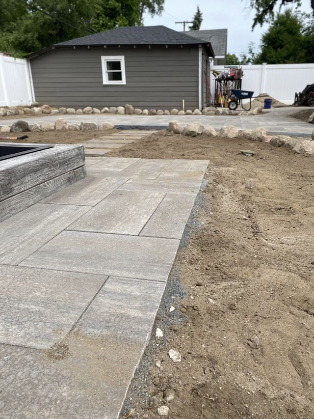 Hardscape installation in progress for a residential paver patio and walkway, showing large rectangular gray pavers being laid next to dirt and a garage.