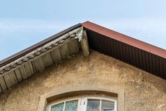 Roofing Restoration Services - Roofing Direct Ptd Ltd A weathered beige stucco gable wall under a blue sky, featuring a damaged wooden eave and dark brown roof trim.