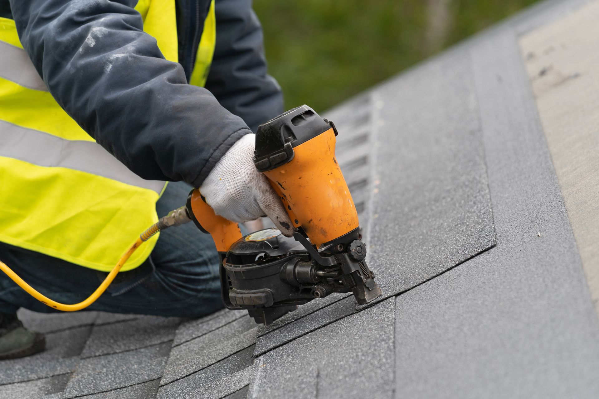 A cropped view of a roofer is using a pneumatic nail gun to install a tile on a roof of a house.