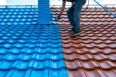 Roofing Painting Services - Roofing Direct Ptd Ltd A person uses a paint sprayer to coat half of a tiled roof in bright blue, contrasting with the remaining red tiles.