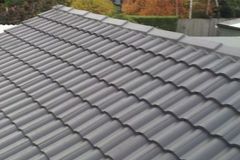 Roofing seal - Roofing Direct Ptd Ltd An angled close-up view of dark gray, wavy roof tiles, with a ridgeline visible against a background of blurred trees.