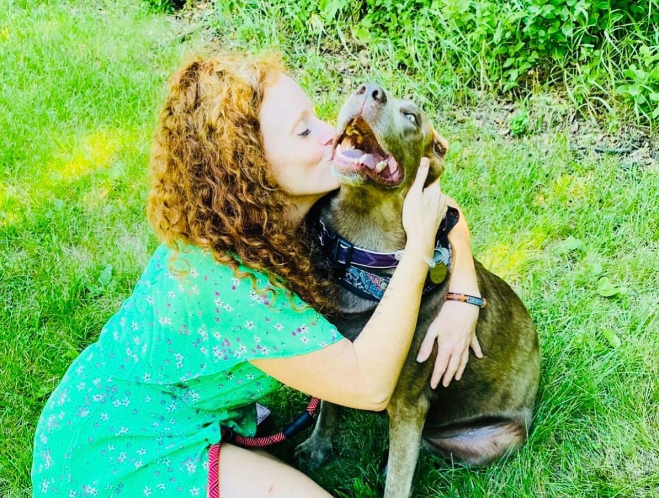 A woman is kissing a dog on the cheek in the grass.