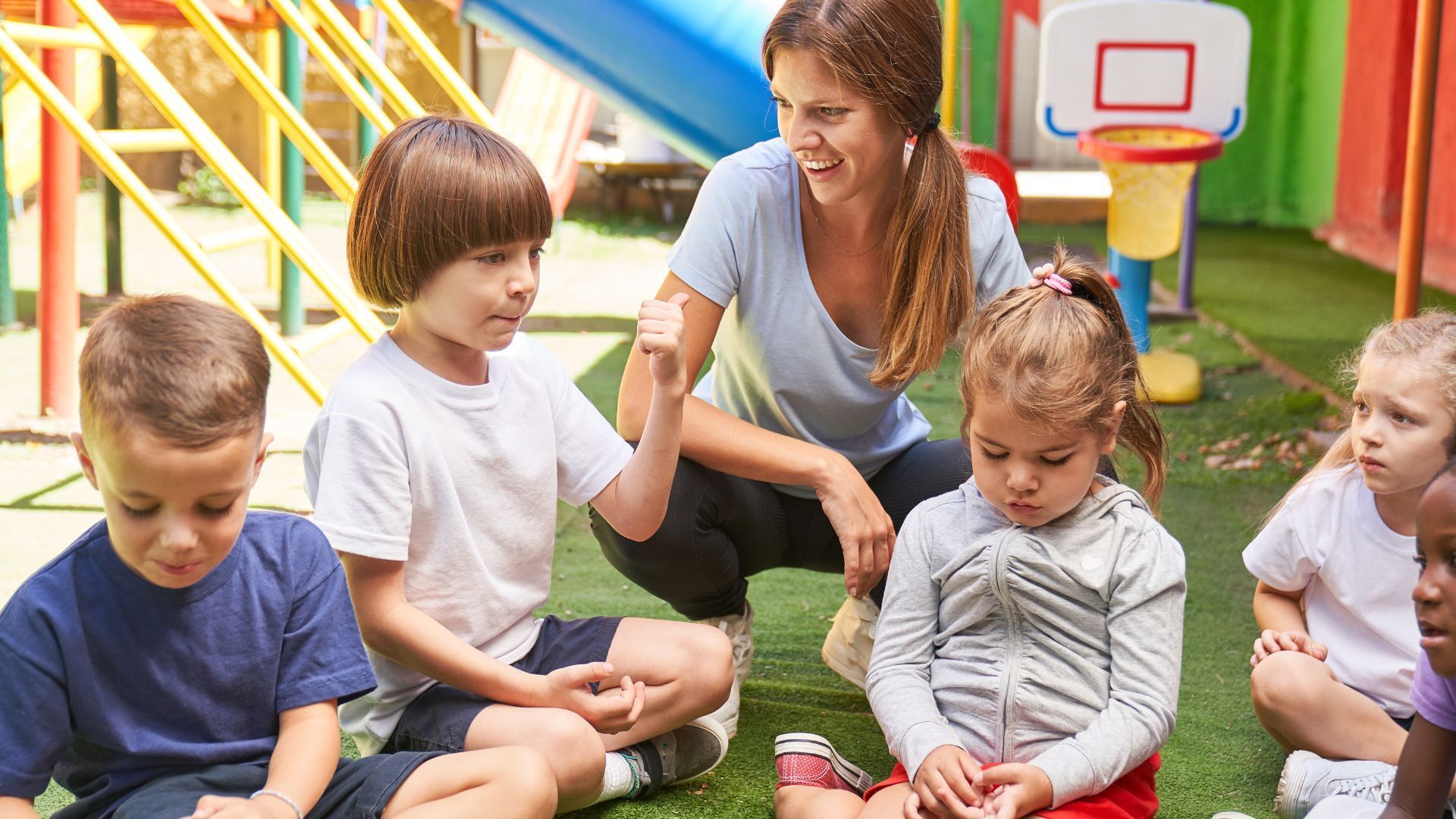 A group of children are sitting on the grass with a teacher.