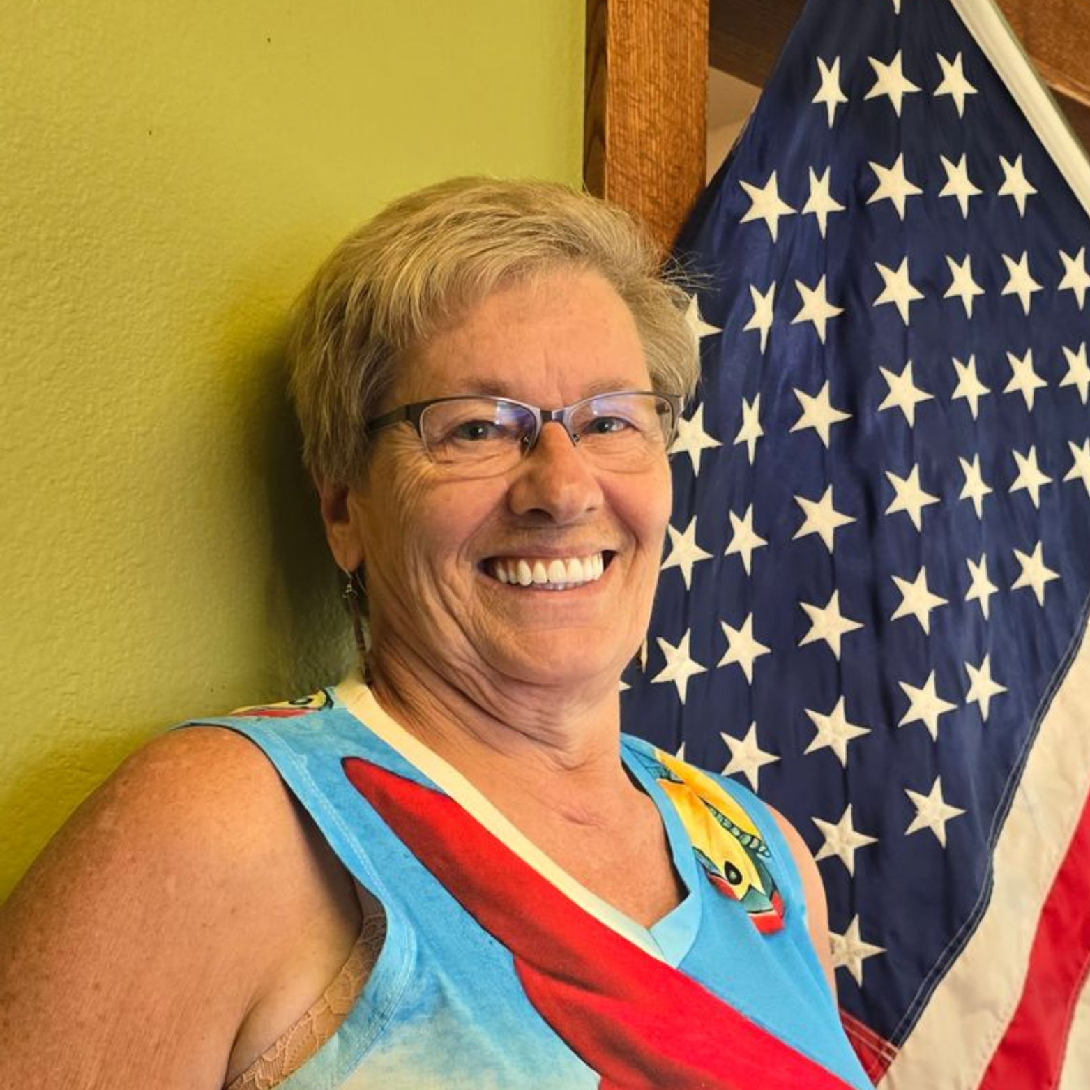 Woman with glasses smiles, next to an American flag. Green wall behind her.