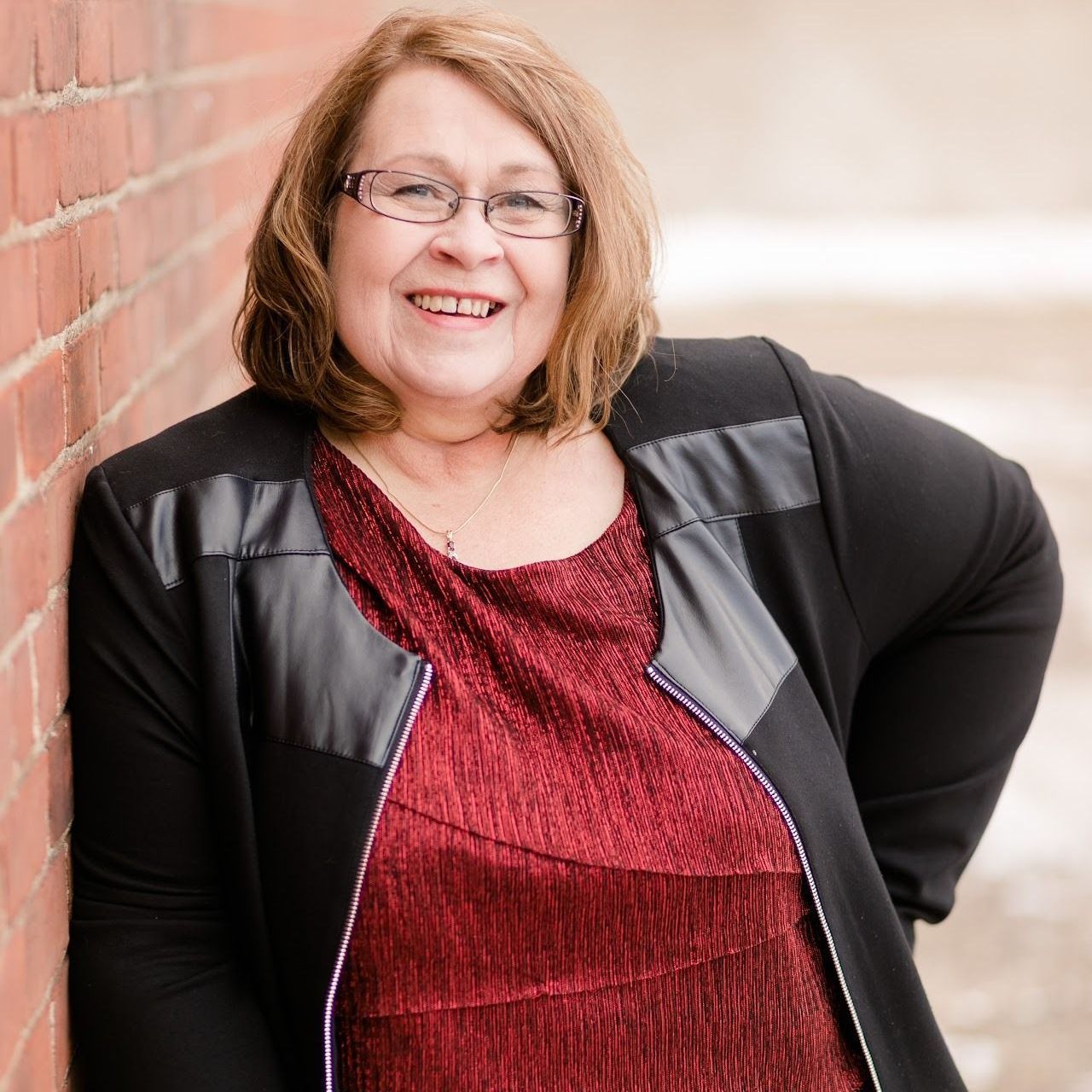 Woman with glasses smiles, leaning against a brick wall; wearing a red top and black jacket.
