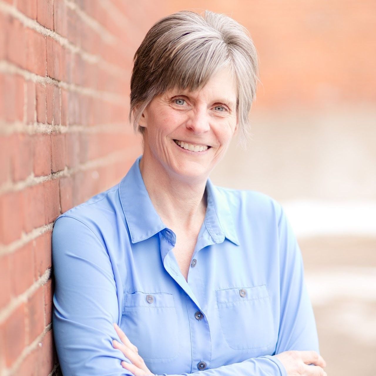 Woman with gray hair, blue shirt, smiling, leaning against a brick wall.