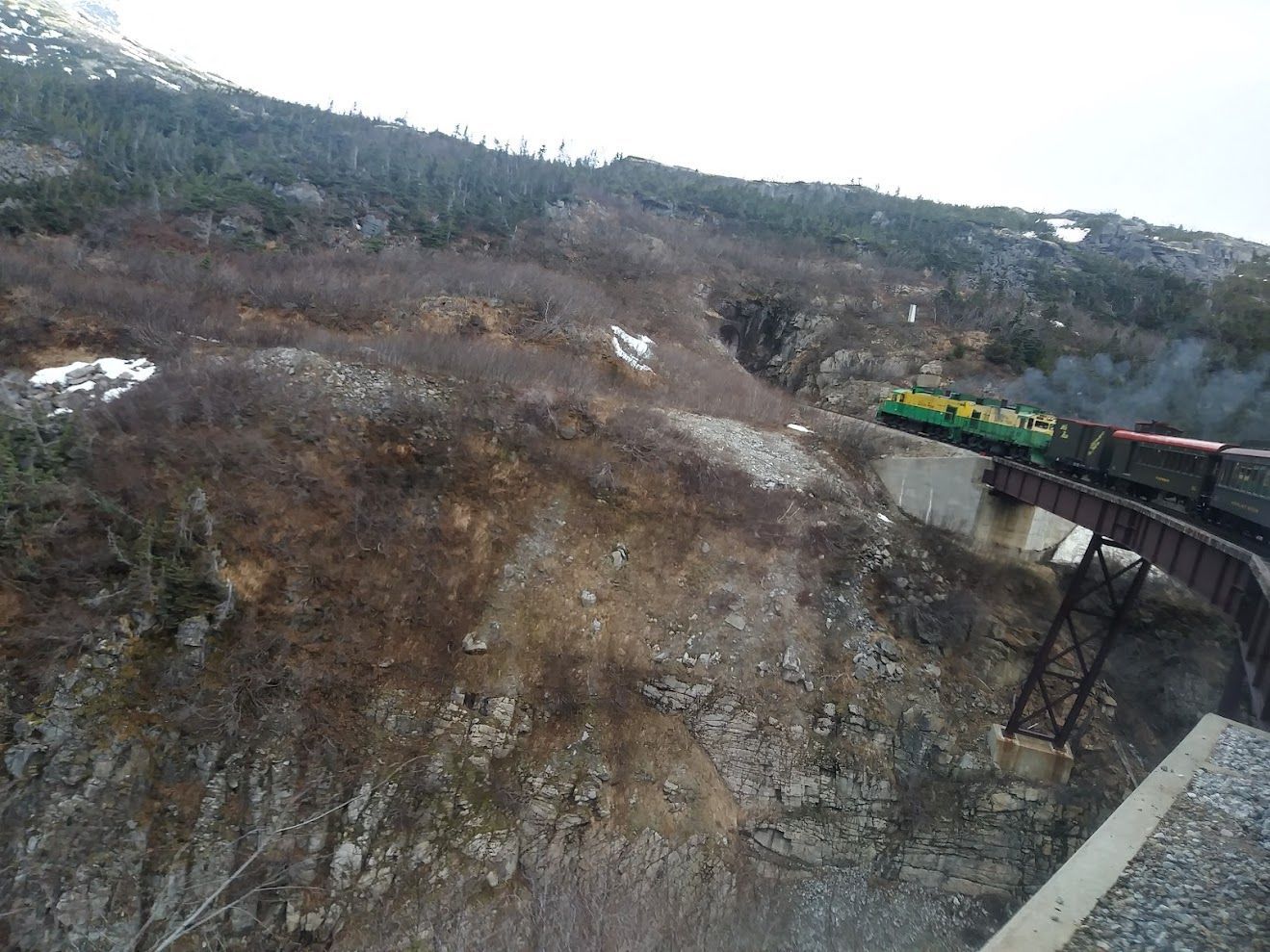 A yellow and green train travels across a high steel trestle bridge over a steep, rocky ravine in a sparse landscape.