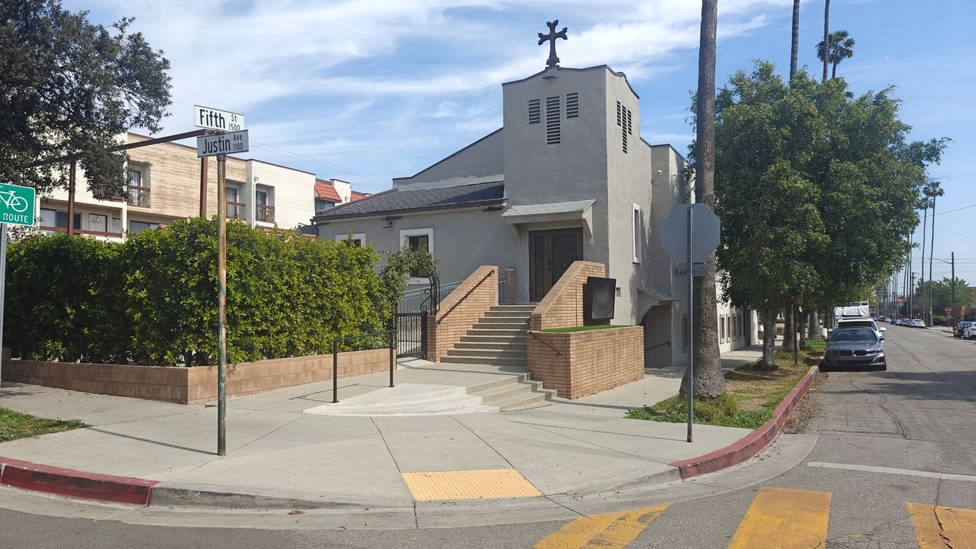 A light-colored church with a cross on top, on a corner with a sidewalk and street, under a blue sky.