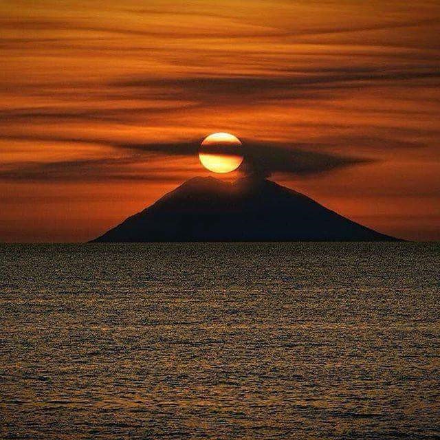 The sun is setting behind a volcano in the middle of the ocean.