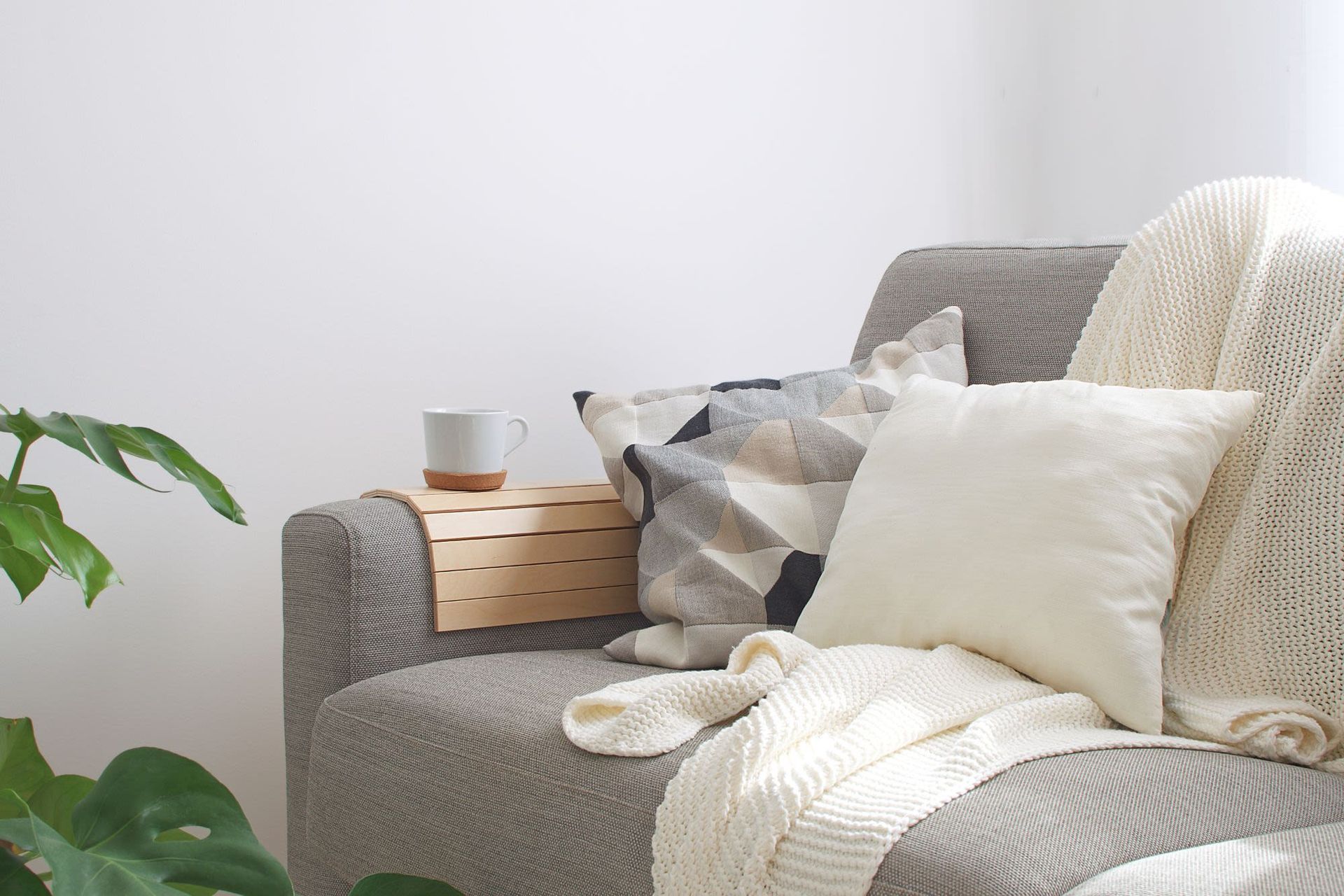 Cozy living room scene with gray sofa, geometric pillow, white knit blanket, and a mug on a wooden armrest tray.