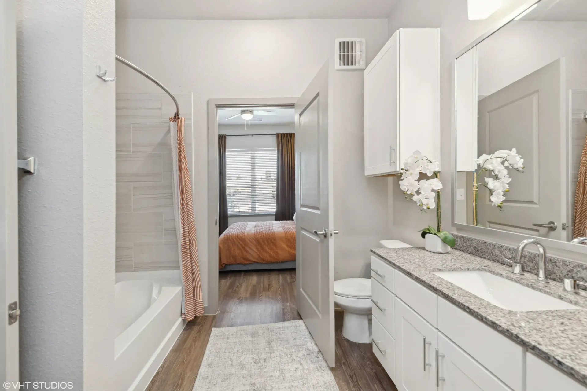 Bathroom with a double-sink vanity, granite countertop, shower/tub, toilet, and doorway to the bedroom.