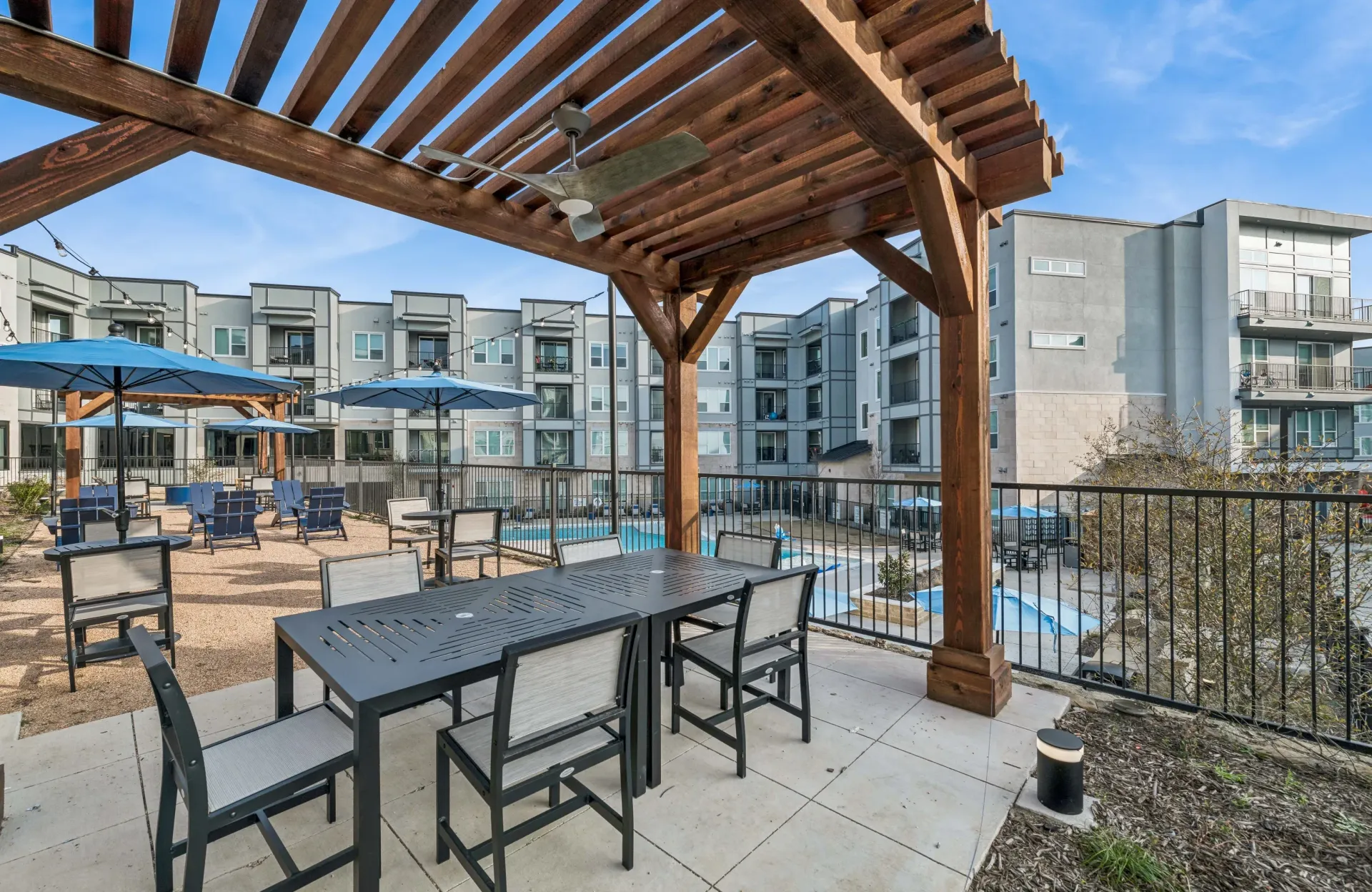 Outdoor community pool area with tables, umbrellas, and a wooden pergola.