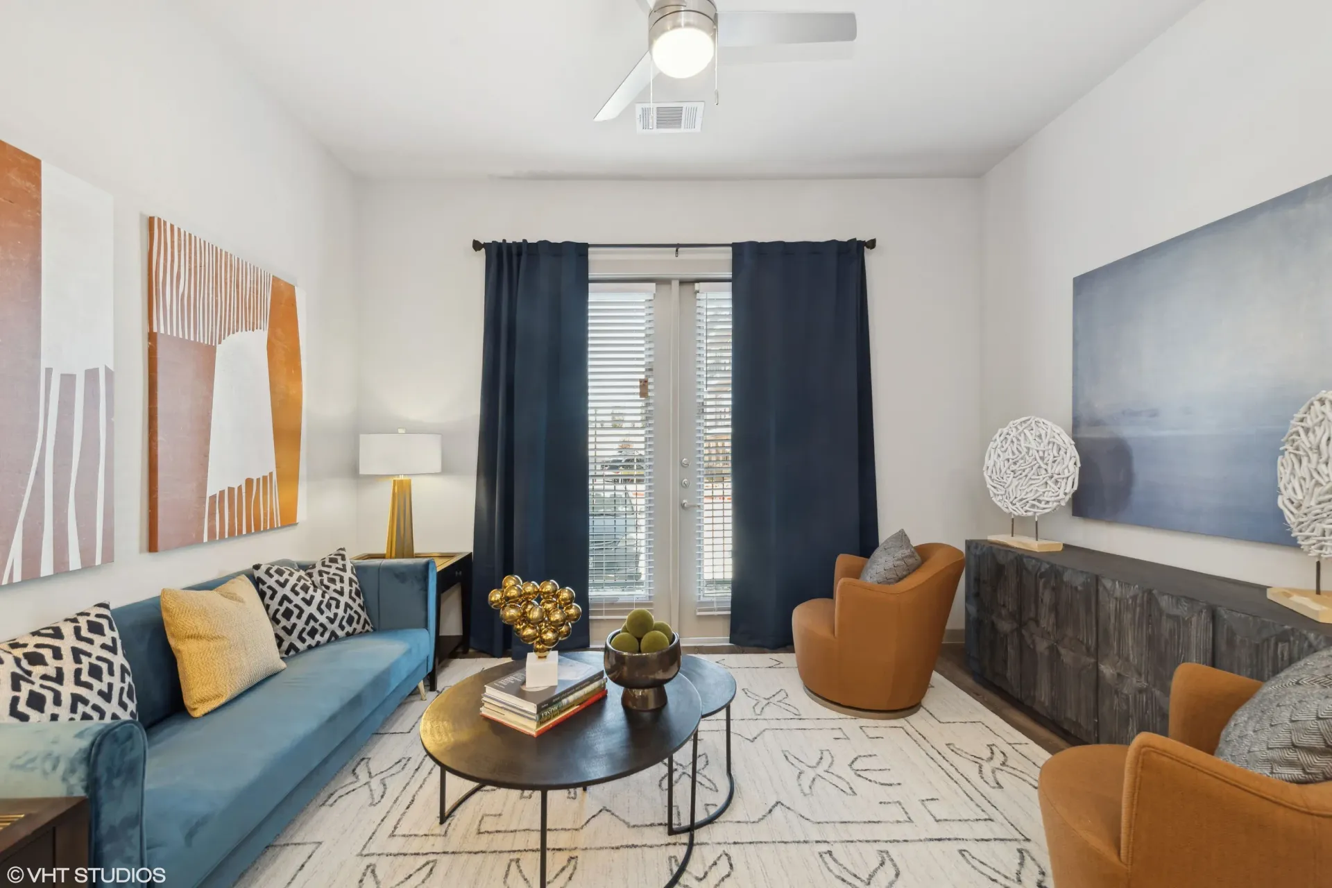 Modern apartment living room with blue sofa, orange chairs, large abstract art, and navy curtains.