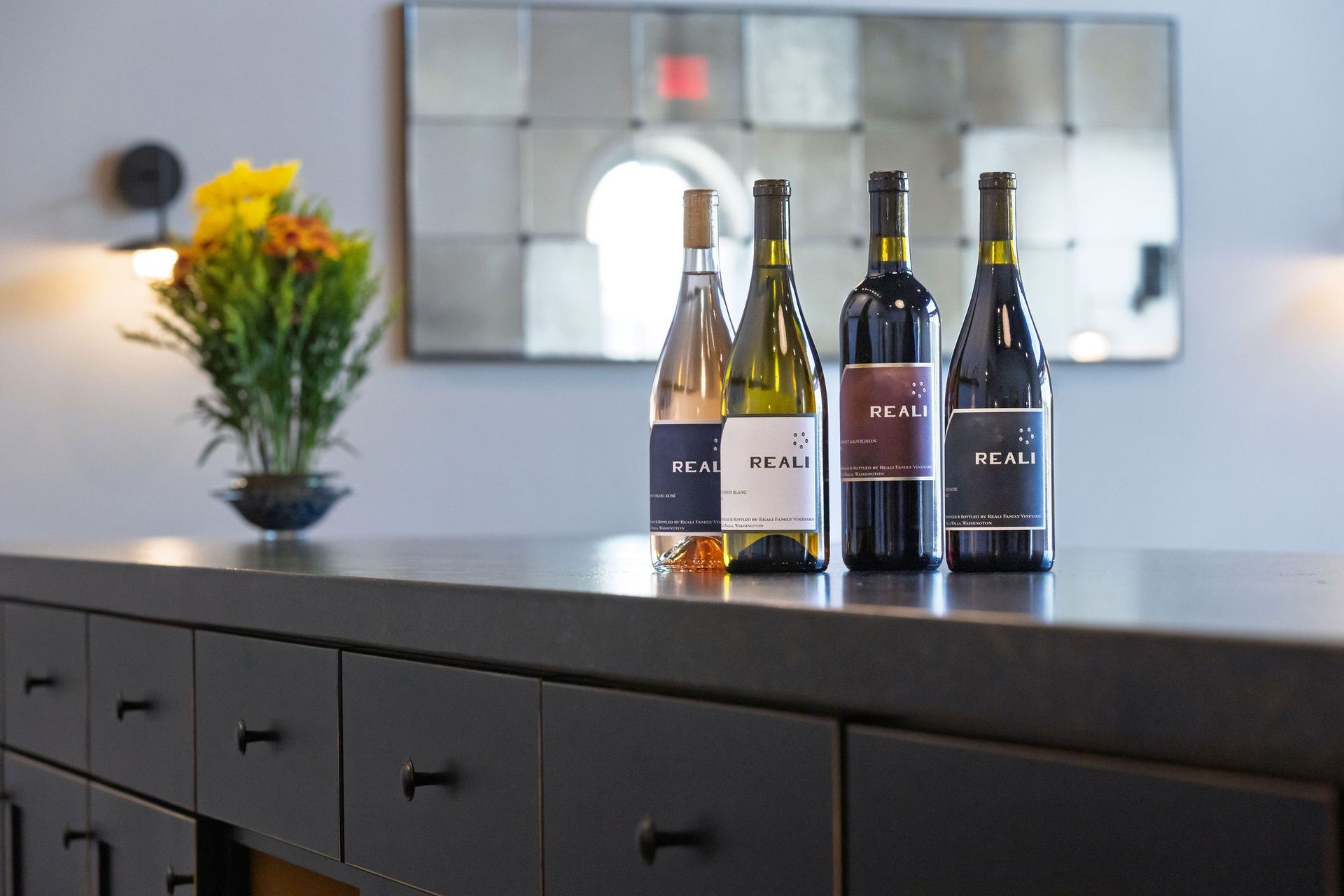 Four bottles of REALI wine on a countertop, with a flower arrangement and mirror in the background.