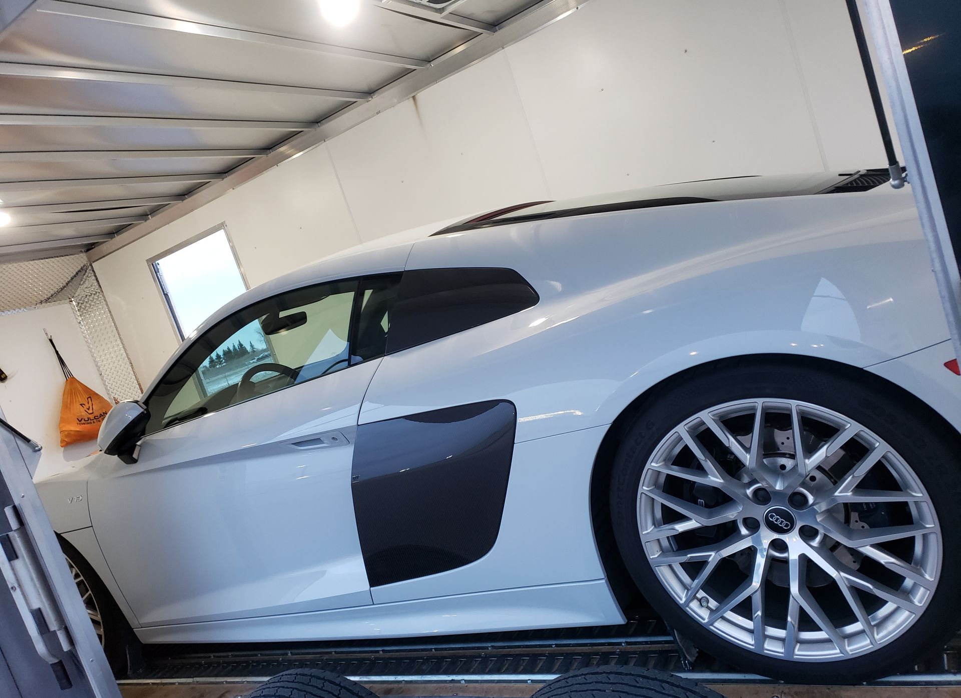 A white sports car is parked in a garage next to a trailer.