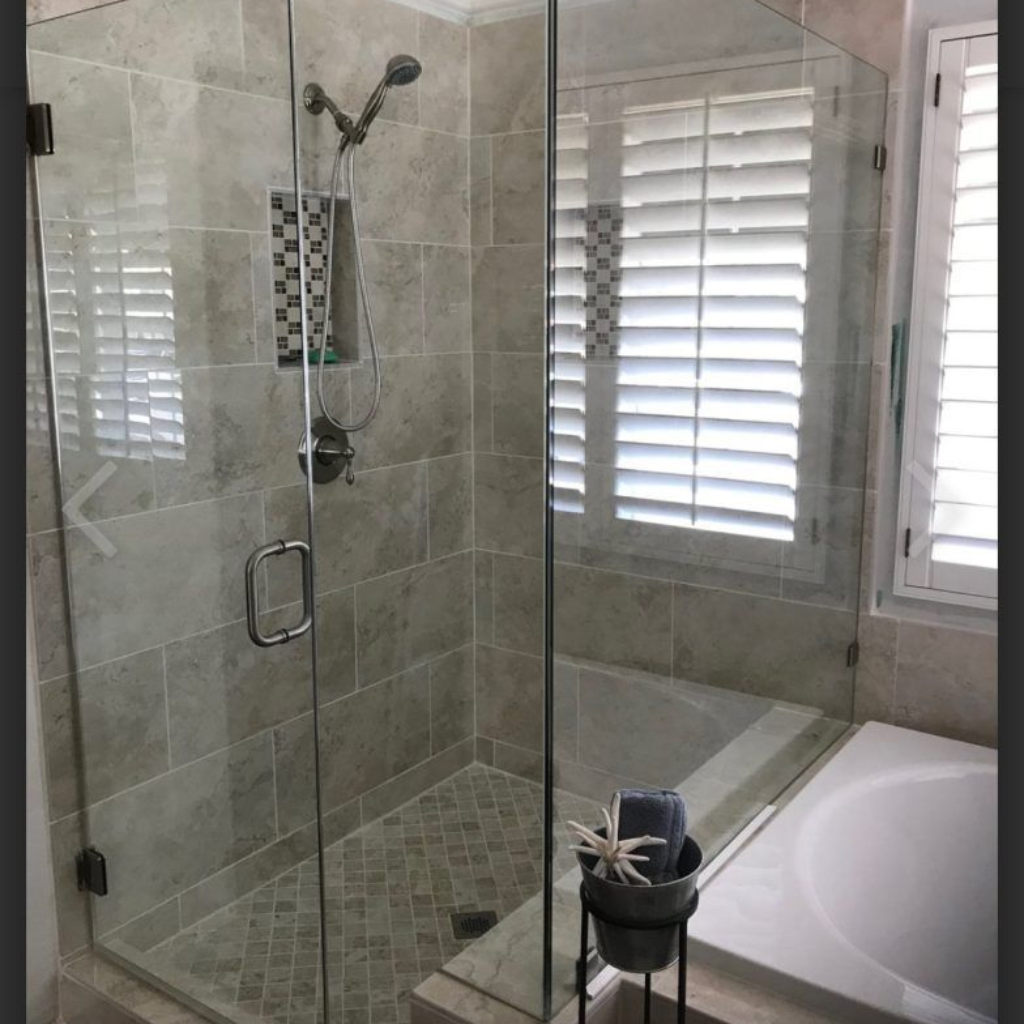 Glass shower with gray tile, stone floor, and window with white shutters.