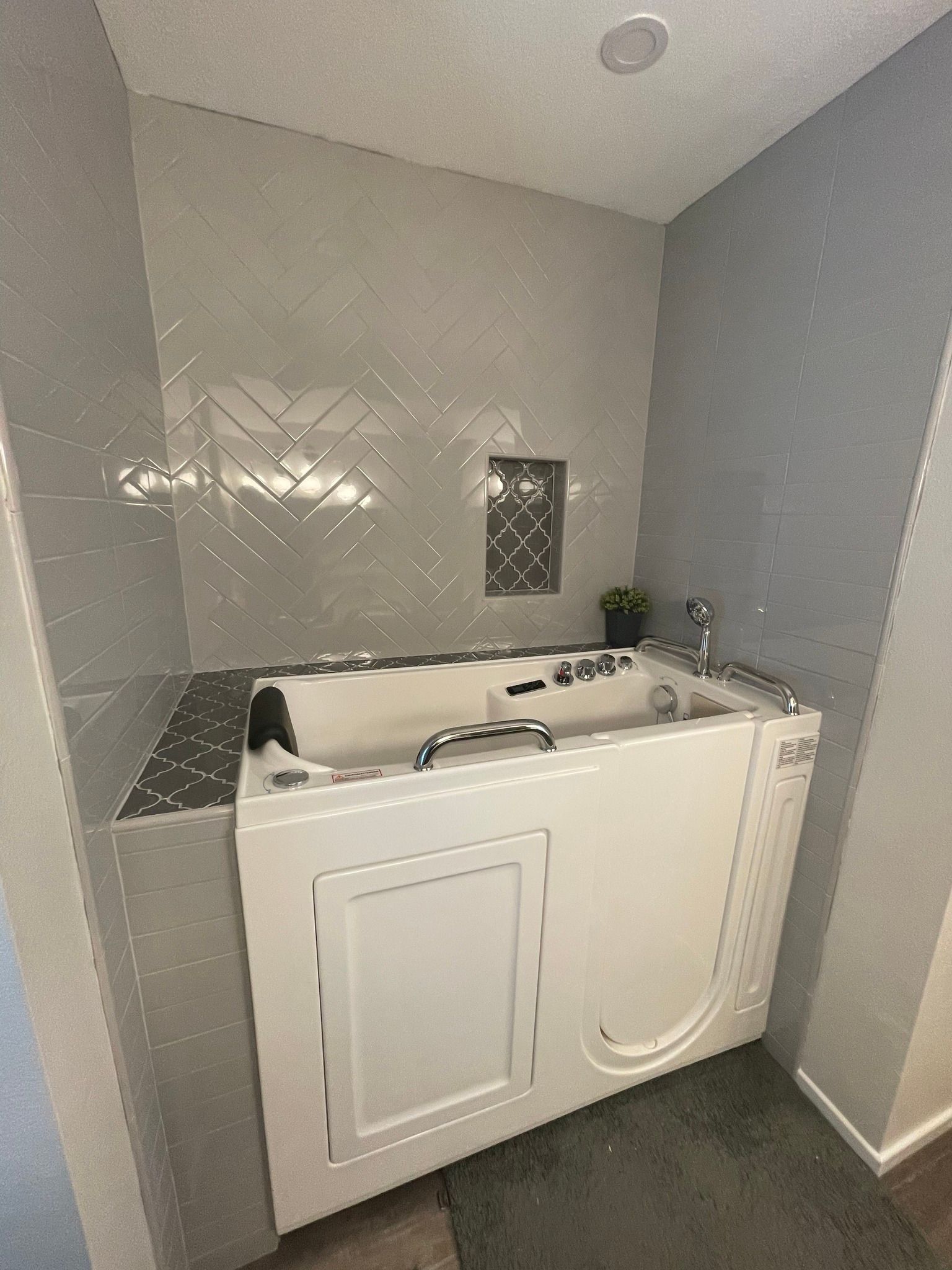 Walk-in bathtub in a gray-tiled alcove. Features include a side door, handrails, and a gray countertop.
