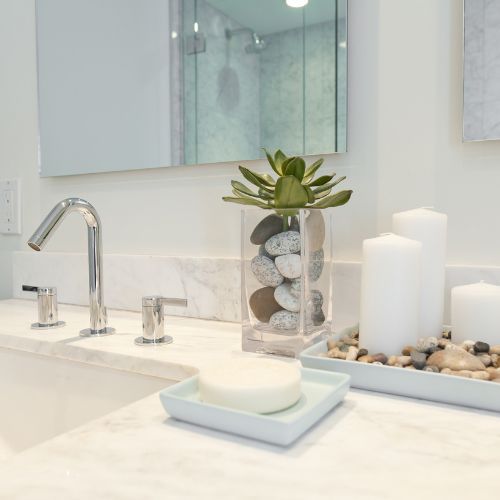 Bathroom countertop with modern faucet, soap, candles, and a plant in a vase with stones.