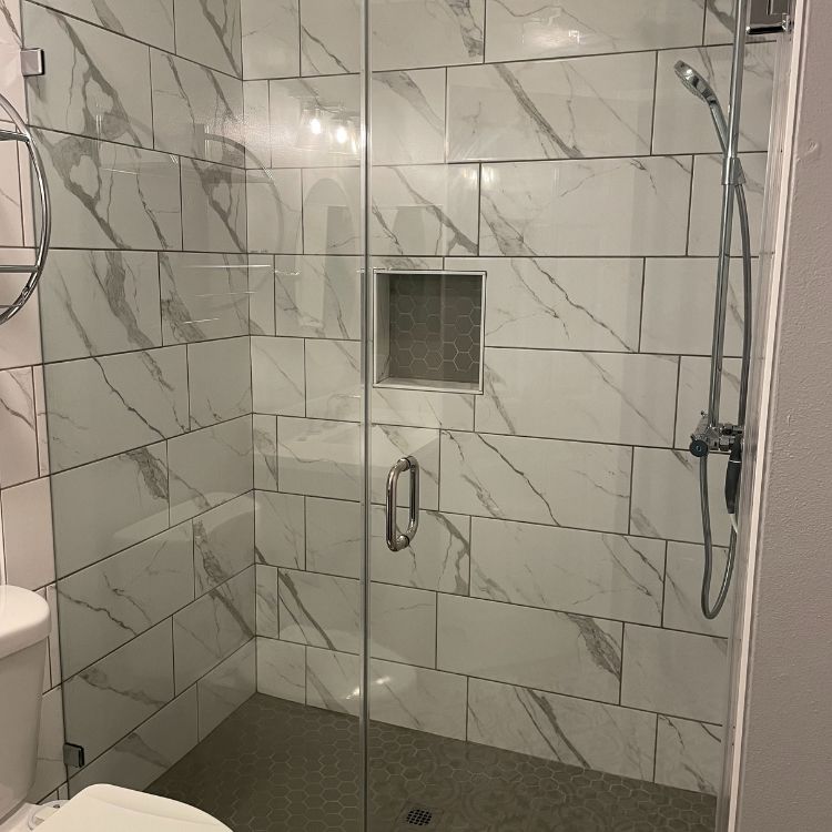 A modern bathroom shower with marble-like tiles and glass doors; a built-in shelf and showerhead are visible.