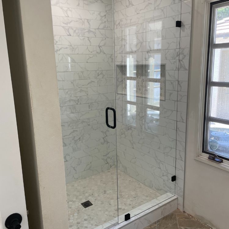 Glass shower enclosure with white marble-look tiles, black hardware, and a small built-in shelf.