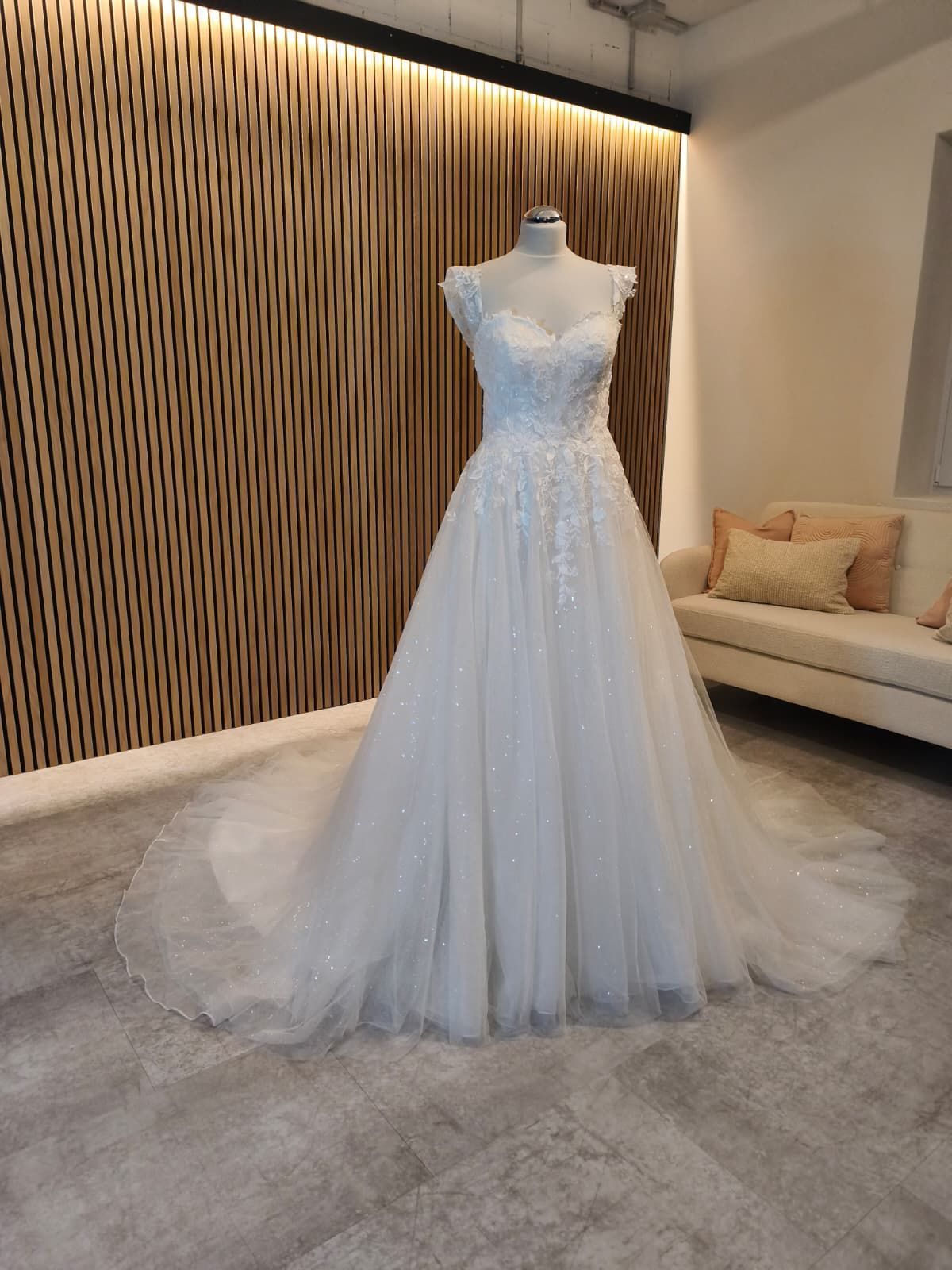 Weißes, perlenbesetztes Brautkleid auf einer Schaufensterpuppe in einem modernen Showroom mit Akzentwand und Sofa aus Holz.