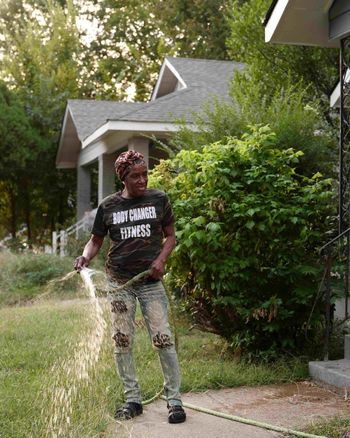 Person spraying water in a driveway beside a house and green shrubs.