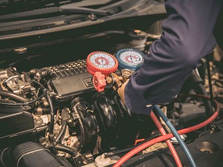 Mechanic repairing car AC with gauges; engine bay in background. Red and blue hoses connect to equipment.