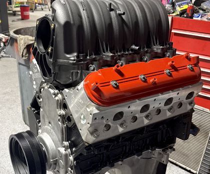 A partially assembled V8 engine with a matte black intake manifold and bright orange valve covers on a shop floor.