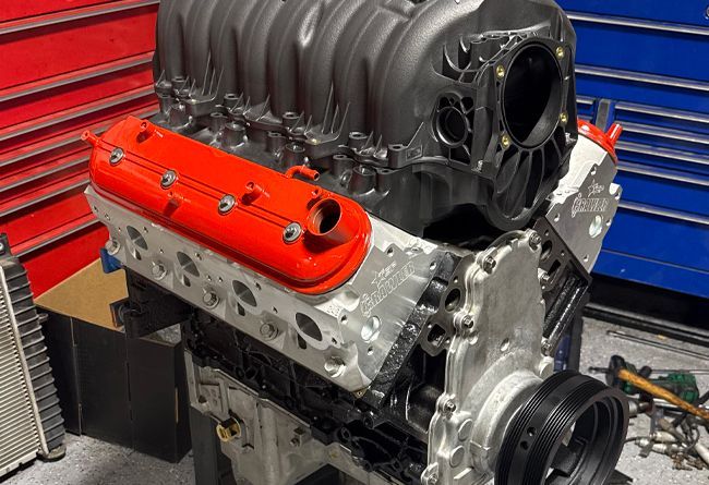 An assembled V8 engine with a black intake manifold and bright orange valve covers, resting in a professional garage.