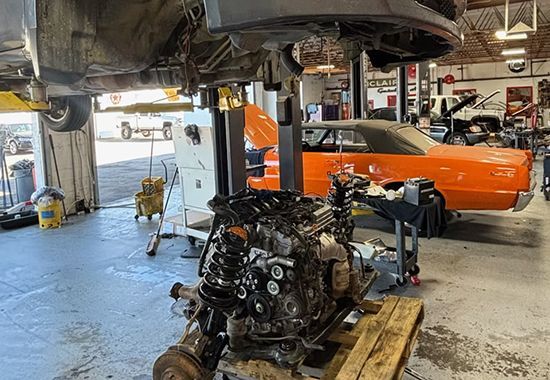 An engine on a wooden pallet in a garage, car on a lift above. Orange classic car in background.