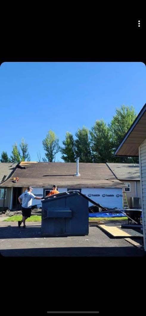 A man is carrying a roof over a dumpster in front of a house.