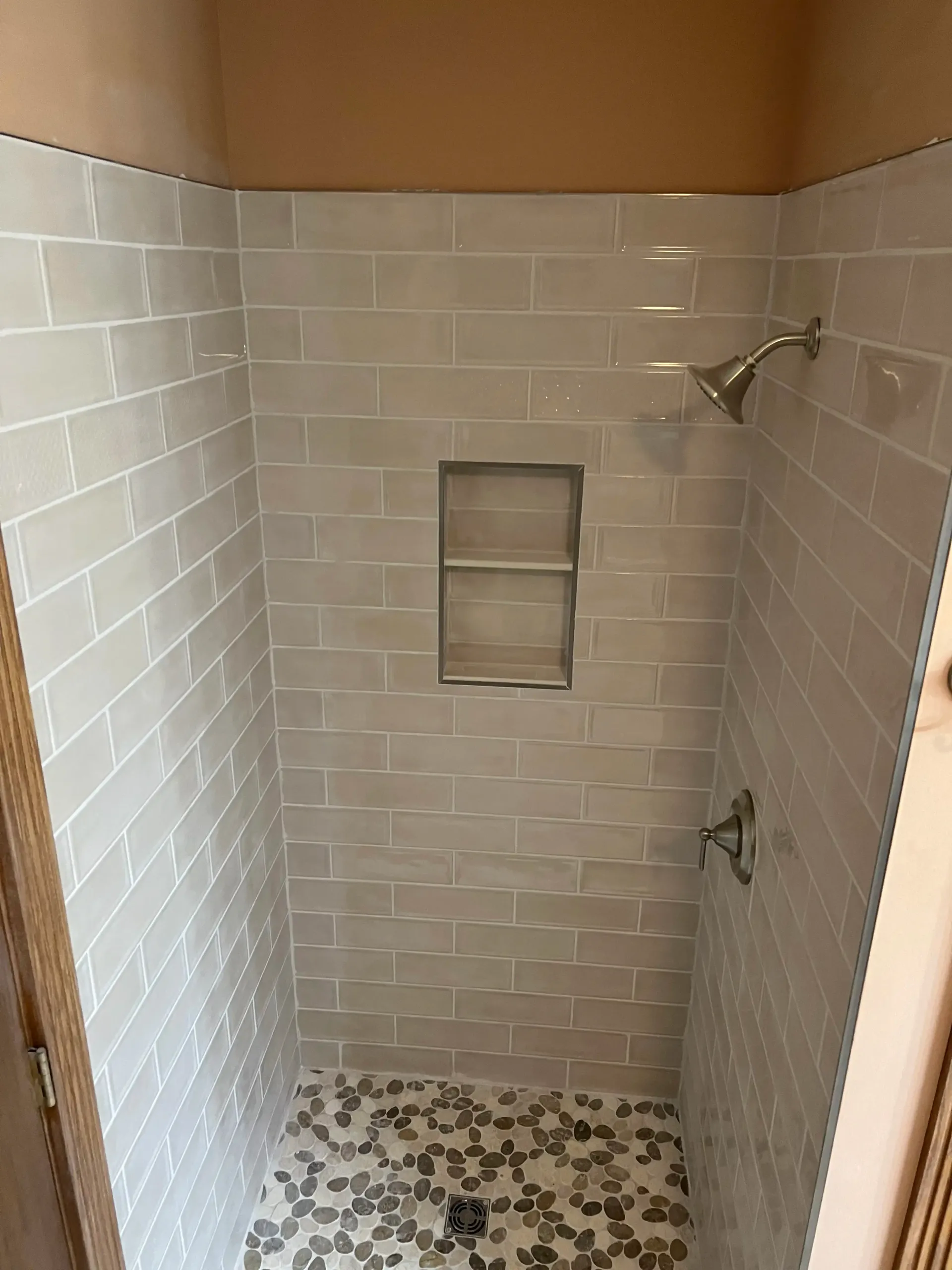 A tiled shower stall with white subway walls, a recessed two-tier shelf, a chrome showerhead, and a stone pebble floor.