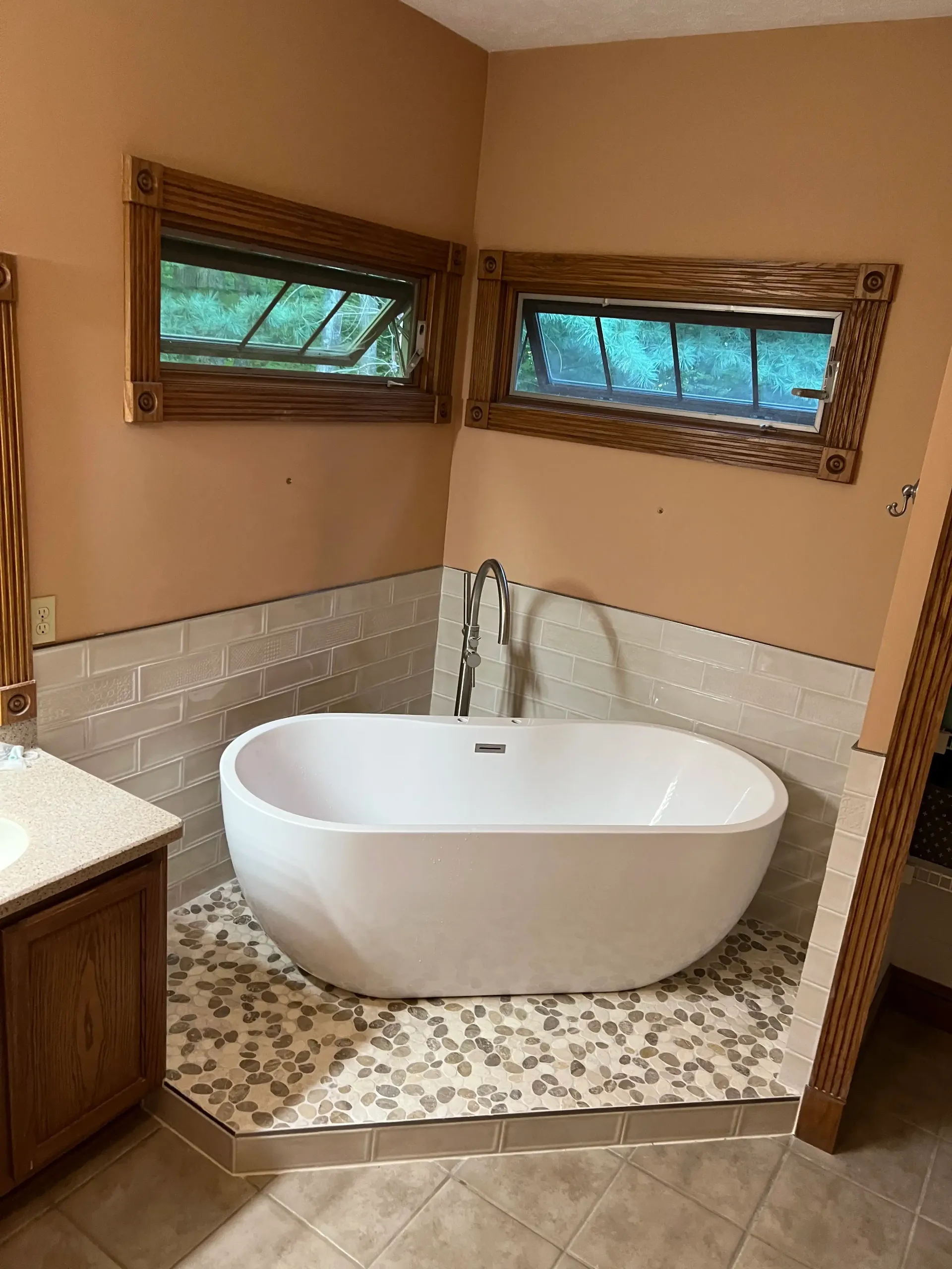 A modern white freestanding bathtub in a tiled corner with two small windows and ornate wooden frames.