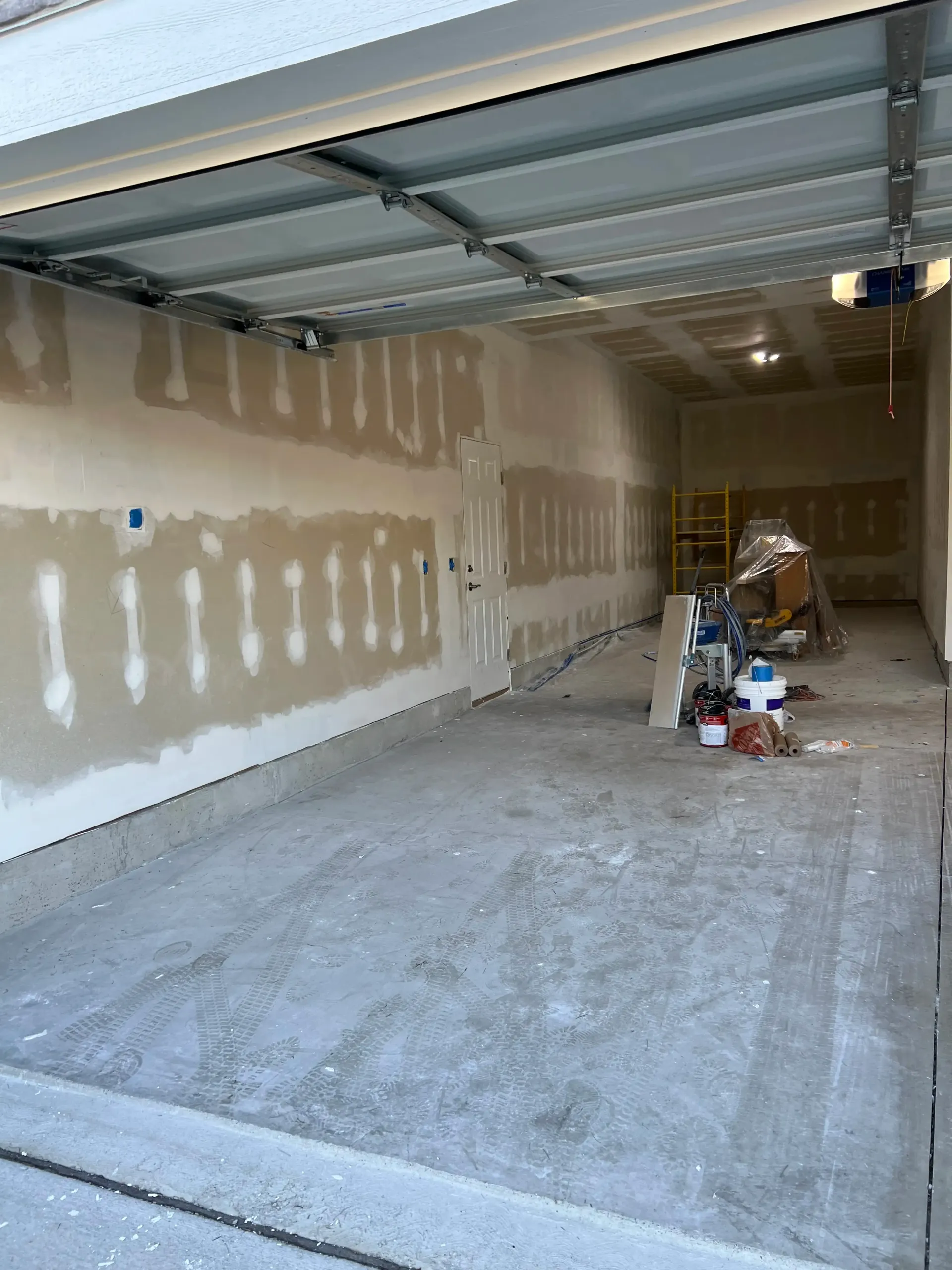 A garage interior under construction with taped and mudded drywall, a concrete floor, and scattered tools and supplies.