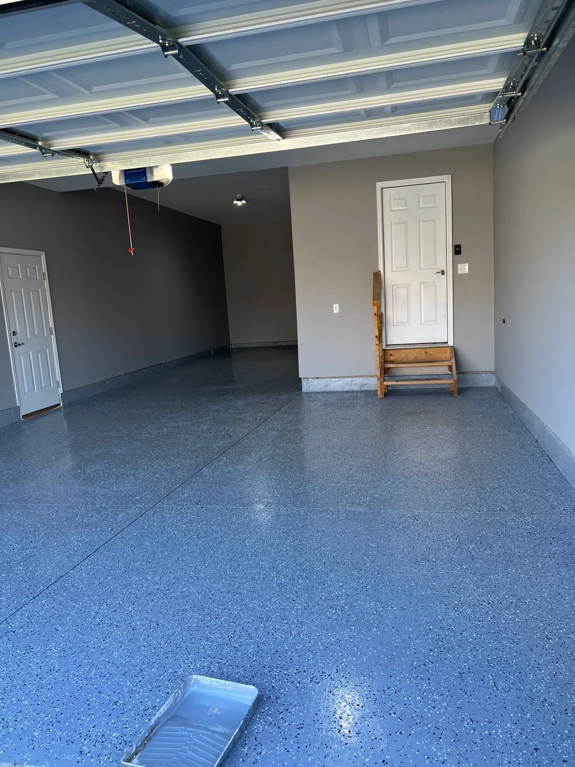A freshly painted gray garage floor with blue and white flecked epoxy, featuring a white door and a small wooden stool.