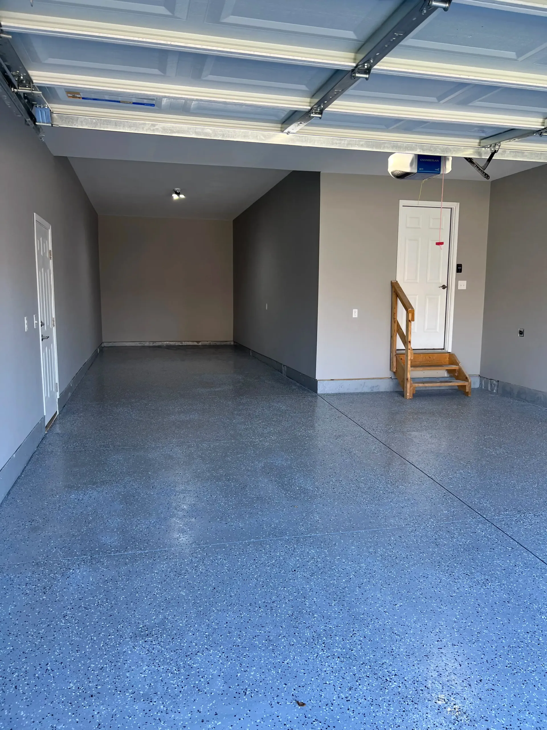 An empty garage with a grey, speckled epoxy floor, white walls, a white door with wooden steps, and an open ceiling.