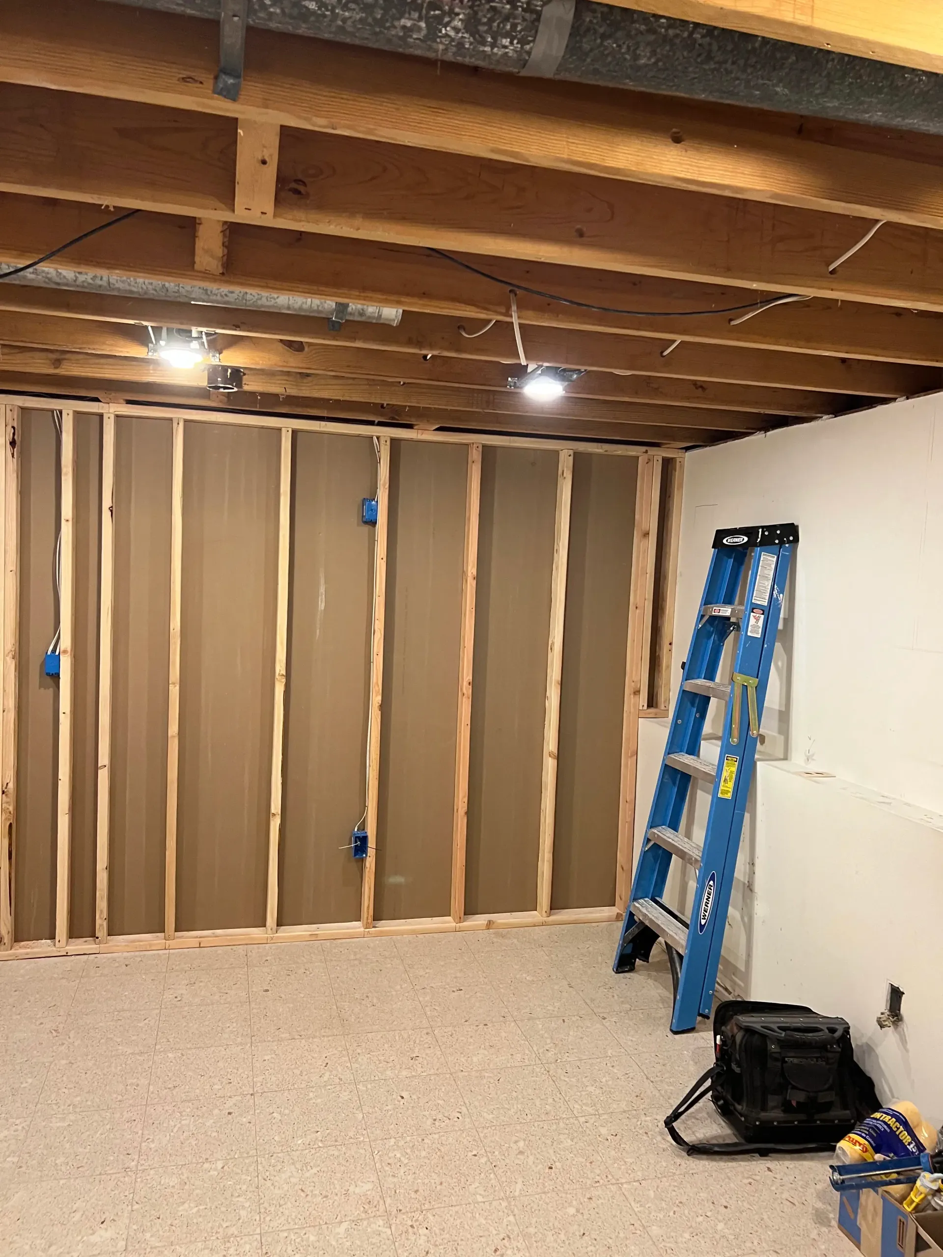 A room under construction with wood-framed walls, exposed ceiling joists, recessed lighting, and a blue ladder.