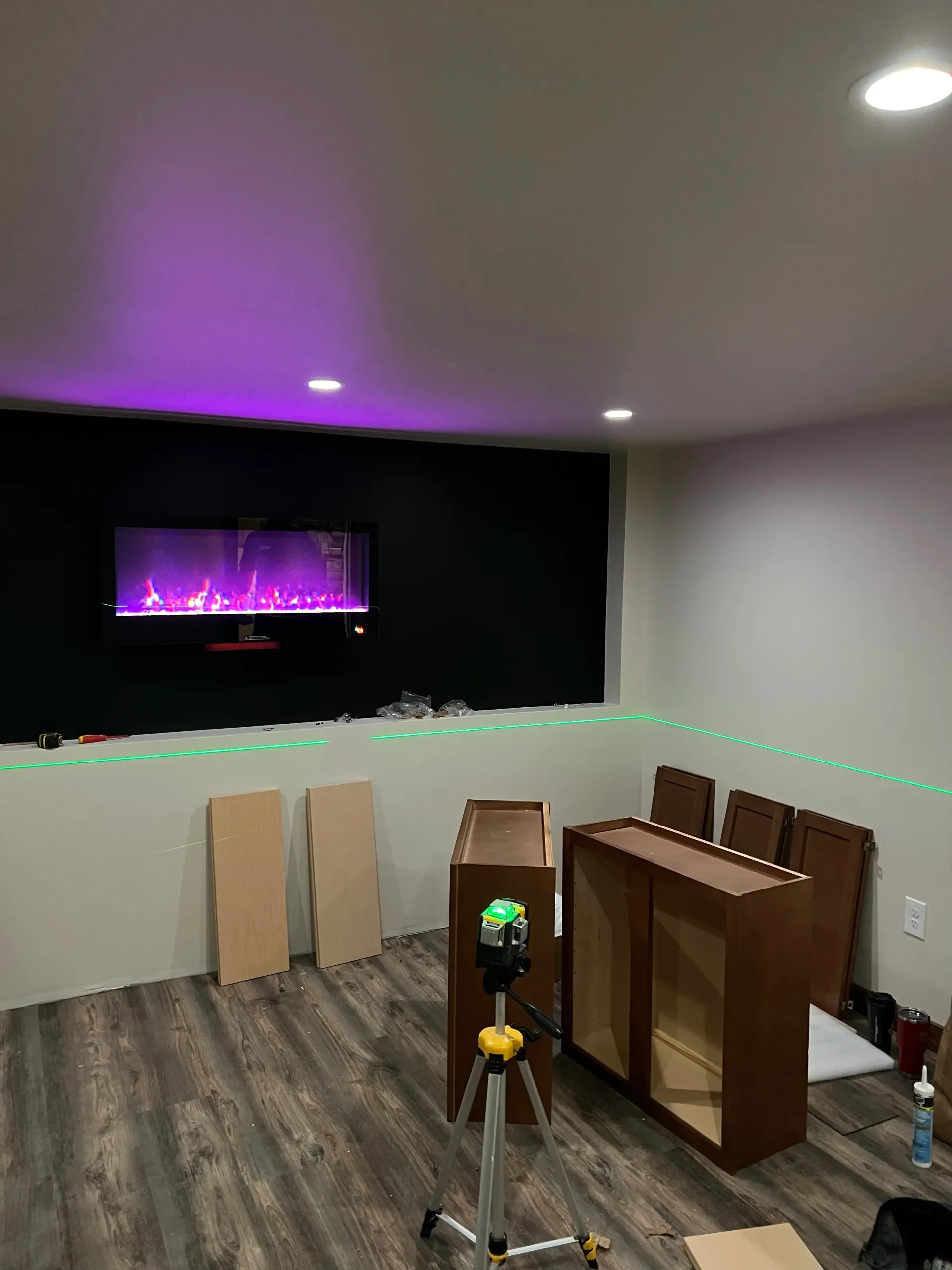 A room under construction with a wall-mounted electric fireplace, a laser level on a tripod, and unassembled cabinets.