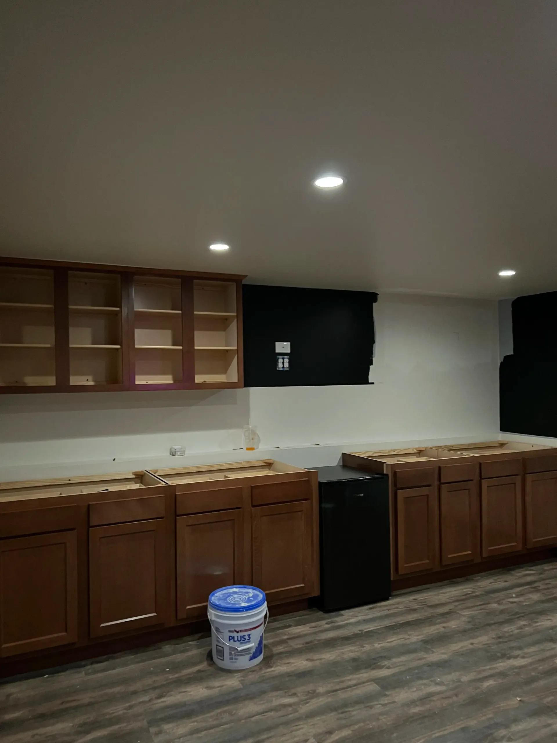 A kitchen undergoing renovation with upper and lower wooden cabinets installed, a black appliance, and a bucket on the floor.