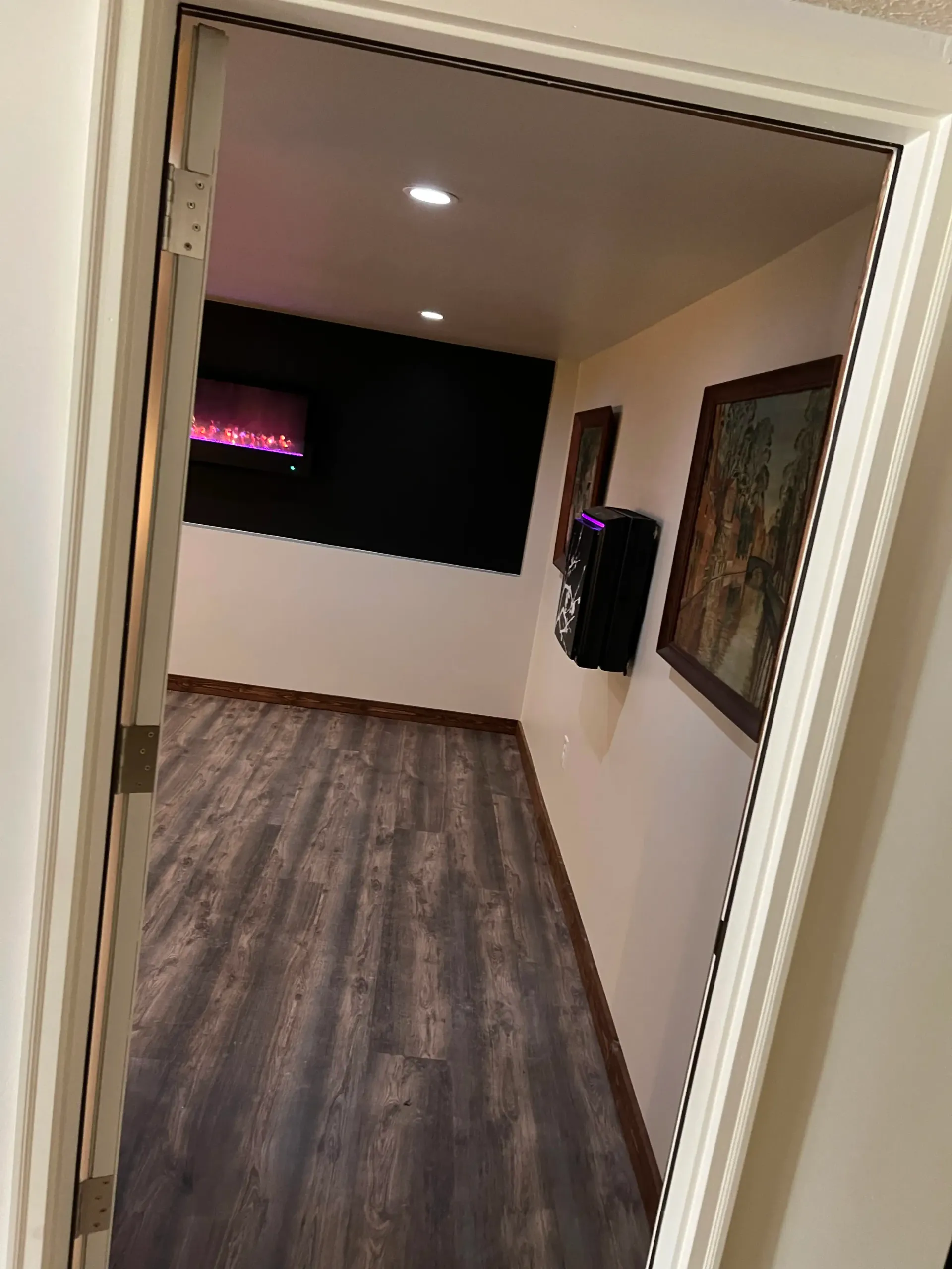 A doorway opens into a room with wood-look flooring, a black feature wall with an electric fireplace, and framed artwork.