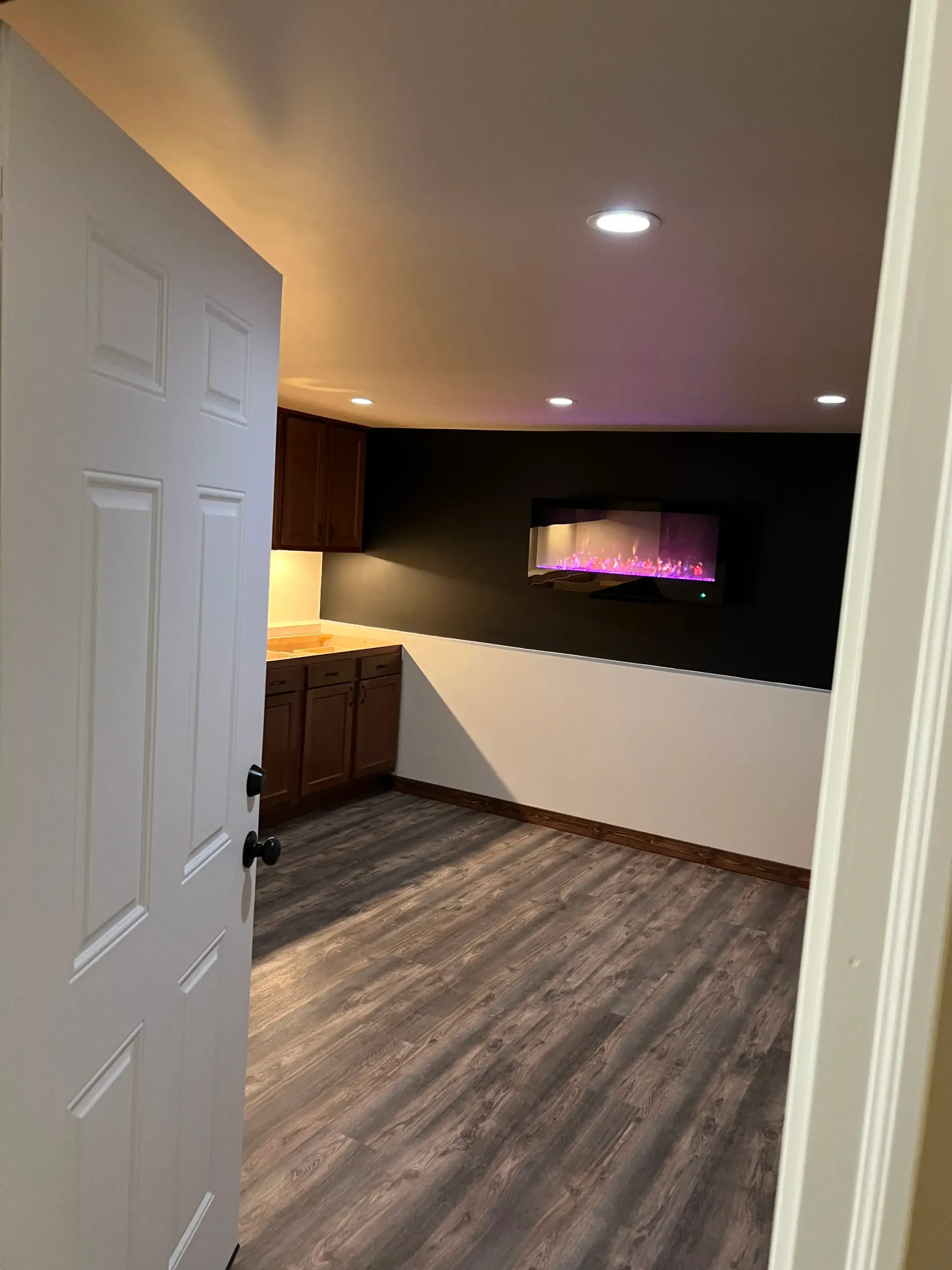A view through an open doorway into a room with gray wood-look flooring, dark cabinetry, and a wall-mounted fireplace.