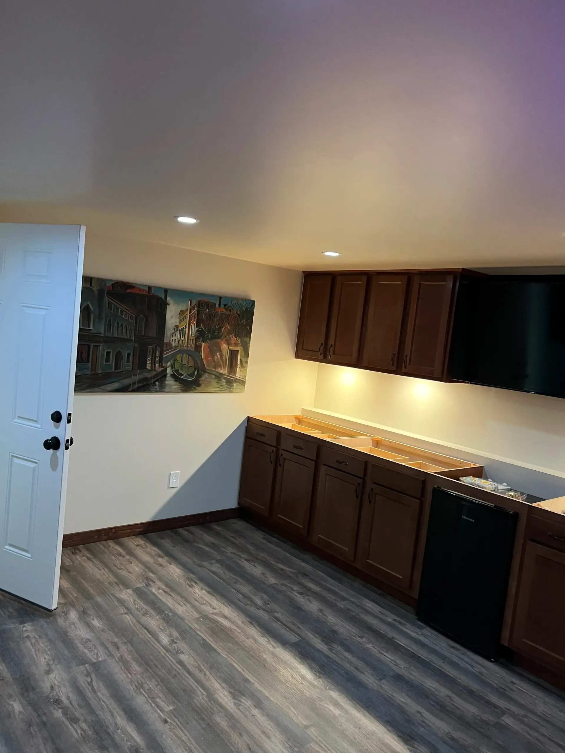 A basement room with dark wood cabinets, a black appliance, and light wood-patterned flooring under recessed lighting.